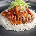 Portrait image of a Chinese chicken and mushroom stir fry served ona black plate with shredded spring onion and white rice