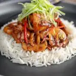 Portrait image of a Chinese chicken and mushroom stir fry served ona black plate with shredded spring onion and white rice