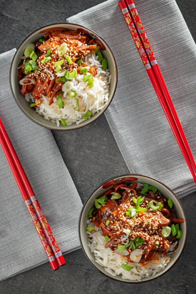 Overhead portrait image of two bowls of chinese orange chicken stir fry served with white rice sprinkled with sesame seeds