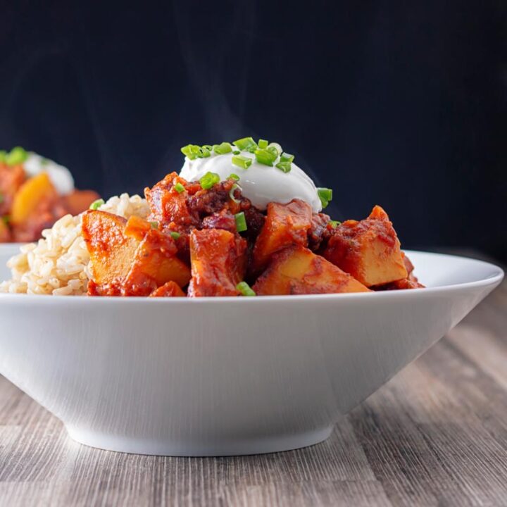 Square image of a steaming hot butternut squash chili served with snipped chives and a dollop of sour cream in a white bowl