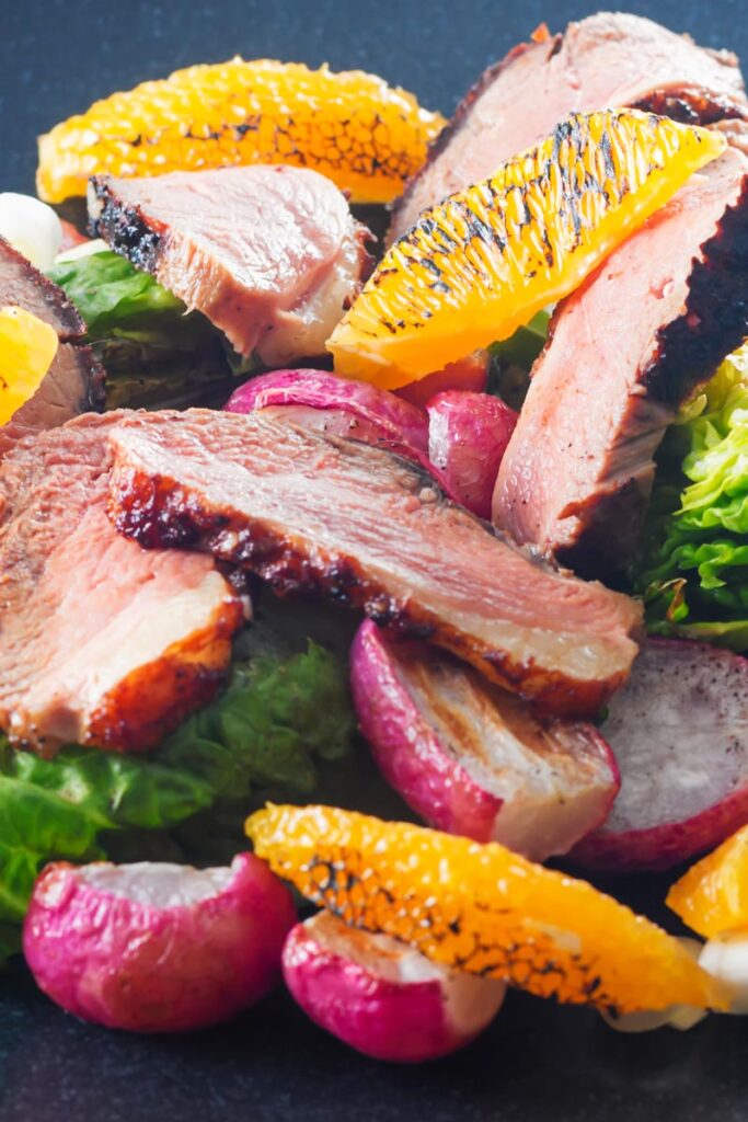 Portrait close up image of a sliced duck breast salad featuring seared orange and roasted radishes served on a black plate