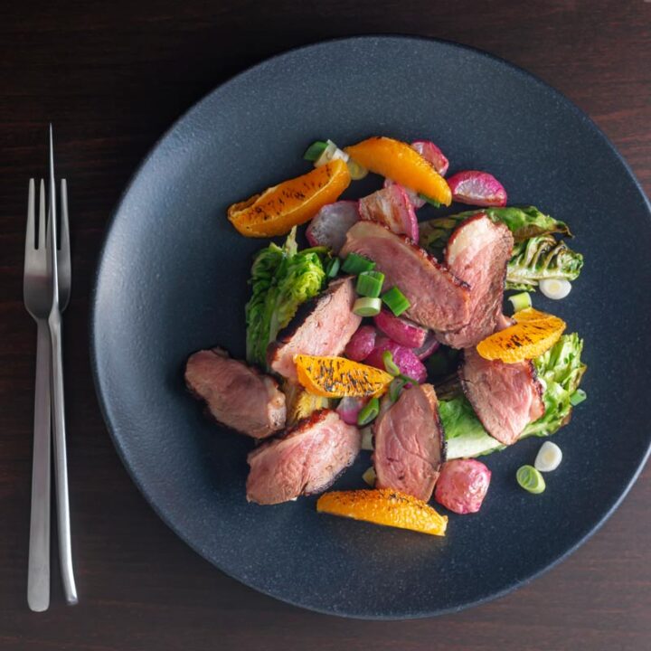 Square overhead image of a sliced duck breast salad featuring seared orange and roasted radishes served on a black plate