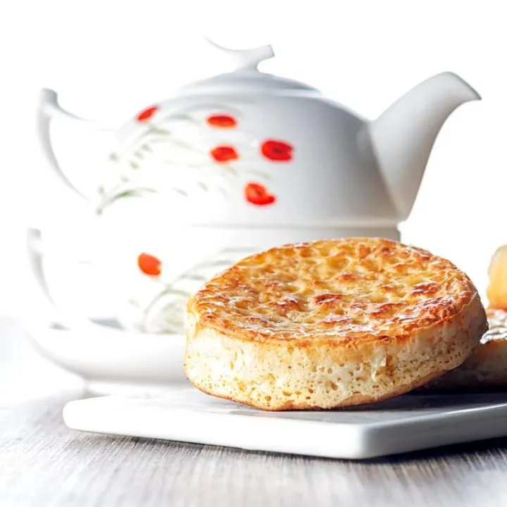 Square image of a toasted homemade English crumpet on a white plate in front of a teapot