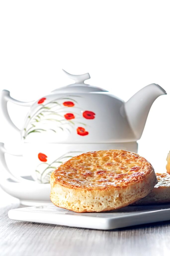 Portrait image of a toasted homemade English crumpet on a white plate in front of a teapot