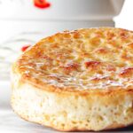 Portrait close up image of a toasted homemade English crumpet on a white plate in front of a teapot