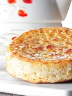 Portrait close up image of a toasted homemade English crumpet on a white plate in front of a teapot