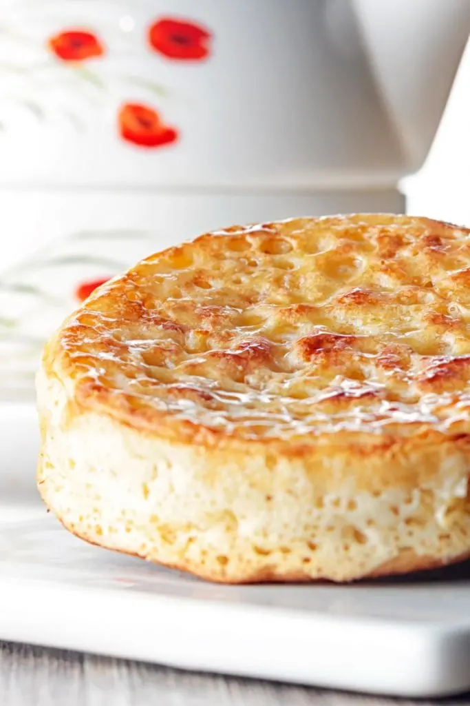 Portrait close up image of a toasted homemade English crumpet on a white plate in front of a teapot