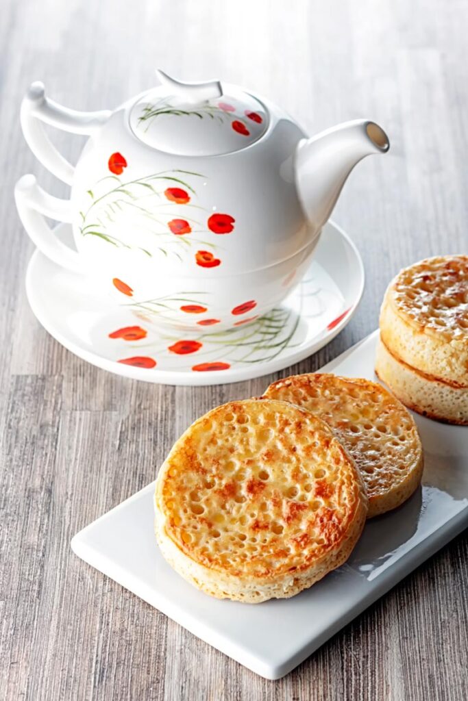 Portrait image of buttered crumpets served on a rectangular white plate with a pot of tea!