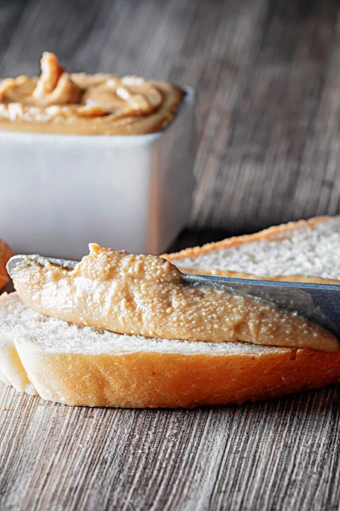 Portrait image of homemade peanut butter being spread on white bread with unshelled peanuts and a bowl of peanut butter in the background