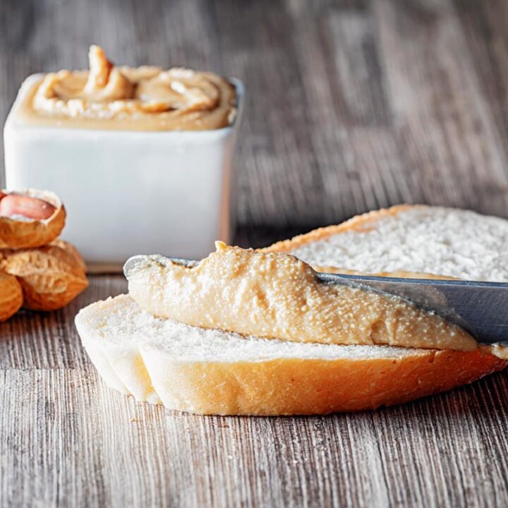 Square image of homemade peanut butter being spread on white bread with unshelled peanuts and a bowl of peanut butter in the background