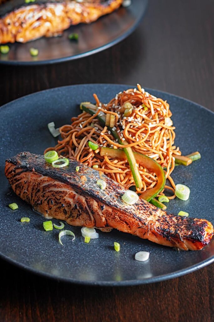 Portrait image of grilled korean salmon fillet with stir fried noodles served on a black plate