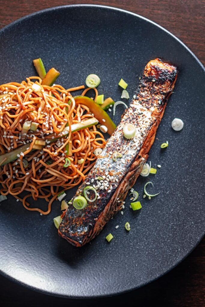 Portrait overhead image of grilled Korean salmon fillet served on a black plate with stir fried noodles