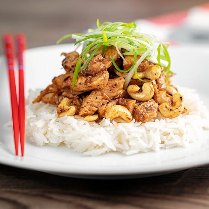 Square image of kung pao chicken served with rice and shredded spring onions on a white pate with red chopsticks