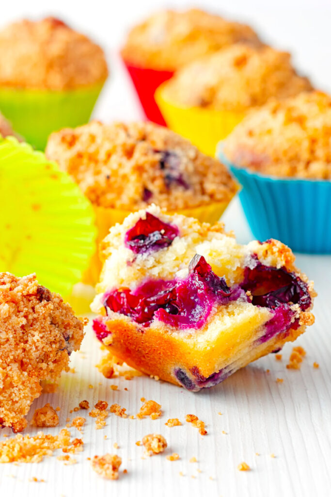Portrait image of lemon and blueberry muffins in colourful silicon muffin cases on a white backdrop with one muffin broken upen showing inside texture