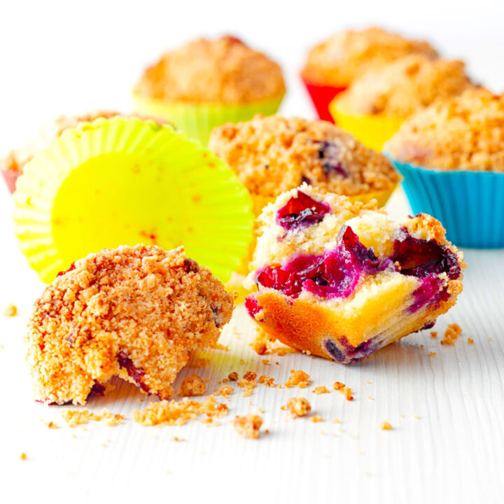 Square image of lemon and blueberry muffins in colourful silicon muffin cases on a white backdrop with one muffin broken open showing inside texture