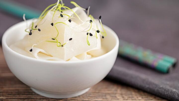 Landscape image of ribbons of pickled daikon served in a small white bowl with sprouted onion seeds