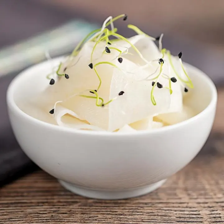 Square image of ribbons of pickled daikon served in a small white bowl with sprouted onion seeds