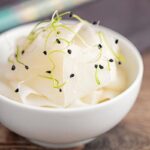 Portrait image of ribbons of pickled daikon served in a small white bowl with sprouted onion seeds