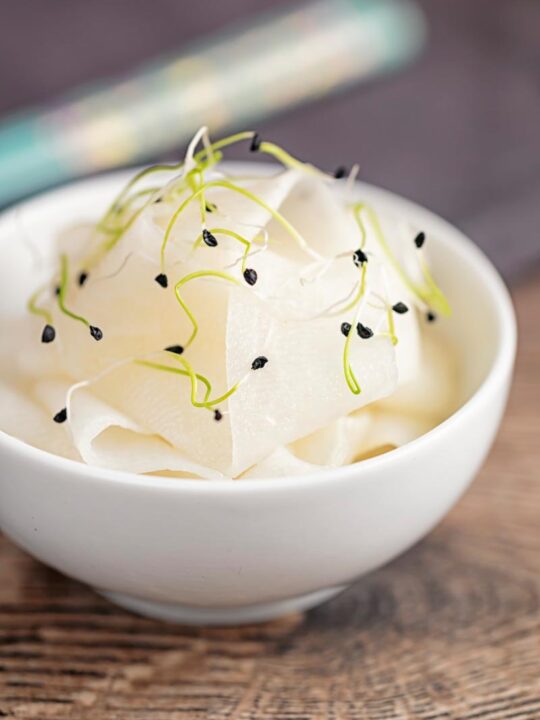 Portrait image of ribbons of pickled daikon served in a small white bowl with sprouted onion seeds