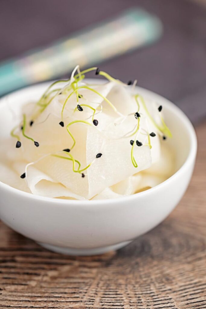 Portrait image of ribbons of pickled daikon served in a small white bowl with sprouted onion seeds