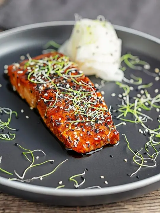 Portrait image of a glazed salmon teriyaki topped with sesame seeds and garnished with sprouted onion seeds served with pickled daikon