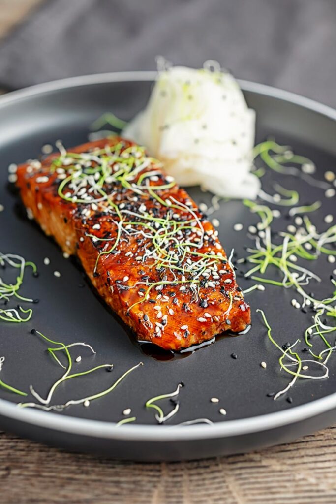 Portrait image of a glazed salmon teriyaki topped with sesame seeds and garnished with sprouted onion seeds served with pickled daikon