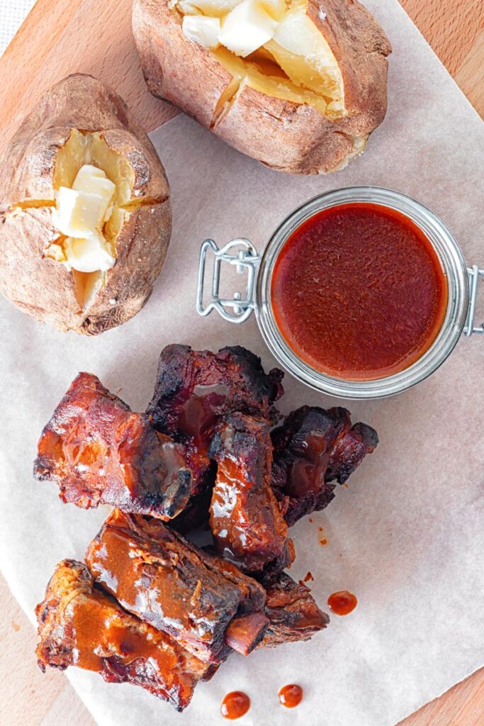 Portrait overhead image of char siu pork ribs that have been cooked in a slow cooker served with baked potatoes and a jar of sauce