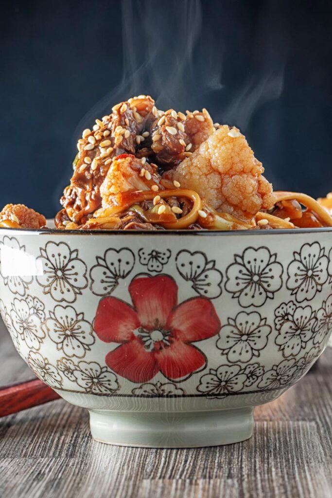 Portrait image of a steaming hot Szechuan beef stir fry with noodles and cauliflower served in an Asian style bowl decorated with a red flower