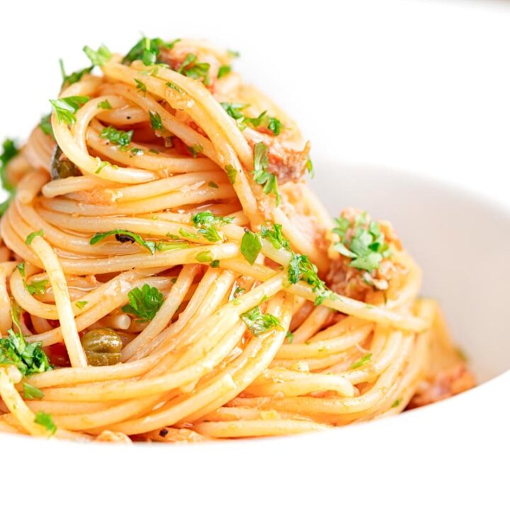 Square close up image of a pile caper, tomato and tuna spaghetti served in a white pasta bowl