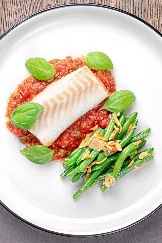 Portrait overhead image of baked cod fillet on tomato sauce with green beans and fresh basil