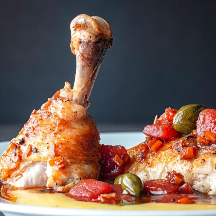 Square image of a chicken leg separated into thigh and drummer with tomatoes and caperberries and a balsamic sauce served on polenta