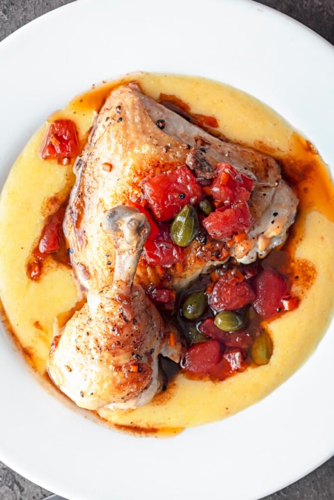 portrait overhead image of a chicken leg separated into thigh and drummer with tomatoes and caperberries and a balsamic sauce served on polenta