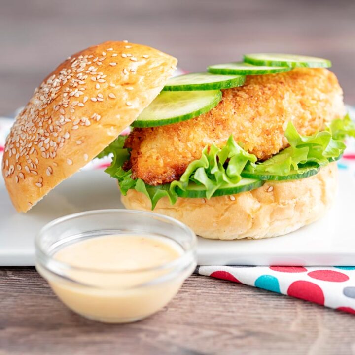 Square image of a chicken breast burger with lettuce and cucumber with a bowl of chilli and lime mayo