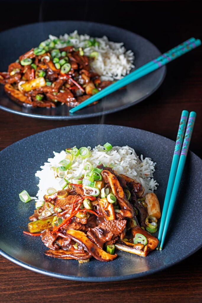 Portrait image of a streaming hot Chinese beef and shiitake mushroom stir fry served on two black plates