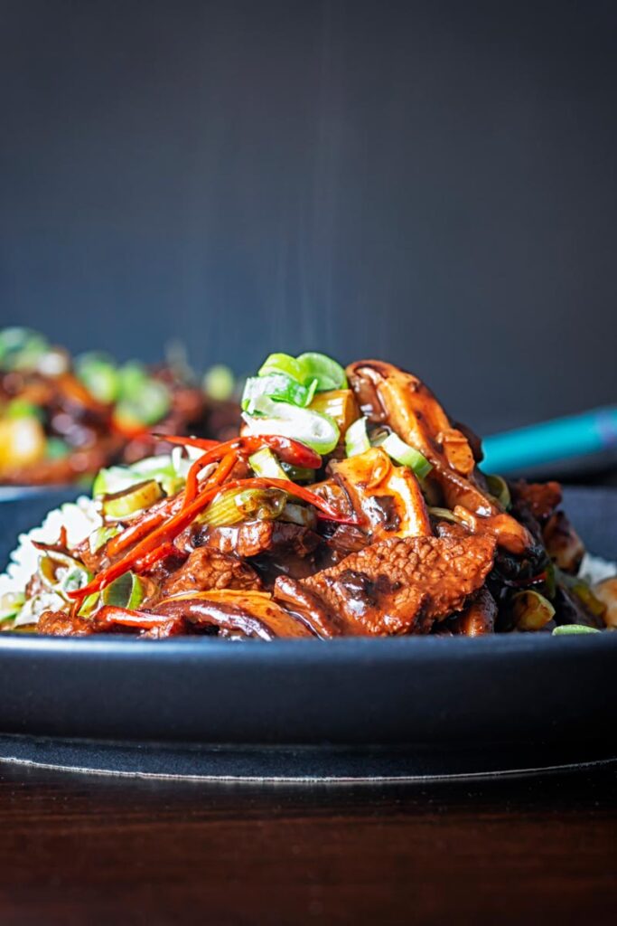 Portrait image of a streaming hot Chinese beef and shiitake mushroom stir fry served on a dark plate