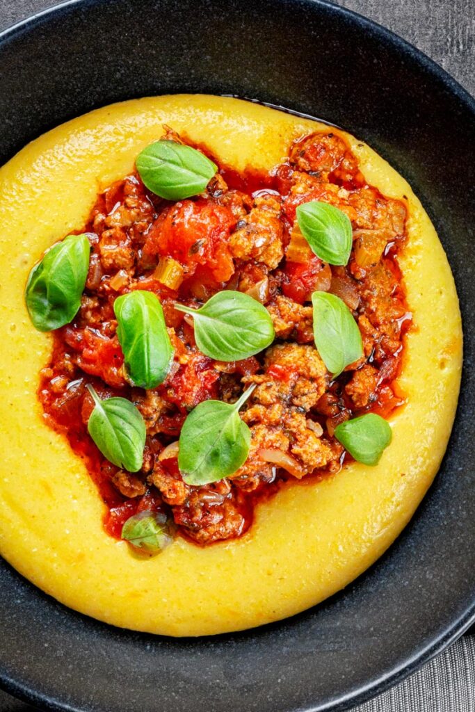 Portrait overhead image of an Italian sausage ragu served on cheesy polenta with fresh basil leaves in a black bowl