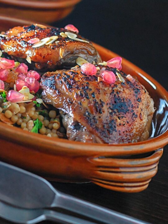 Portrait close up image of pomegranate molasses glazed chicken thighs served with Israeli couscous and pomegranate seeds
