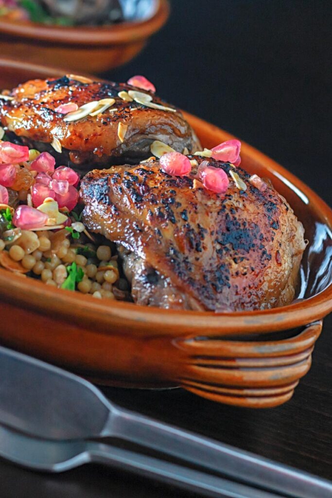 Portrait close up image of pomegranate molasses glazed chicken thighs served with Israeli couscous and pomegranate seeds