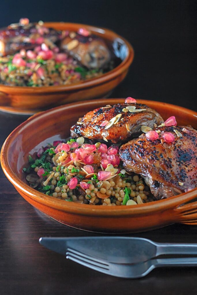 Portrait image of pomegranate molasses glazed chicken thighs served with Israeli couscous and pomegranate seeds