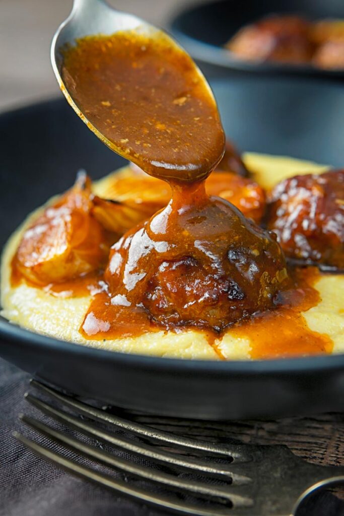 Portrait image of BBQ sauce being poured over pork meatballs served in black plate on a polenta porridge