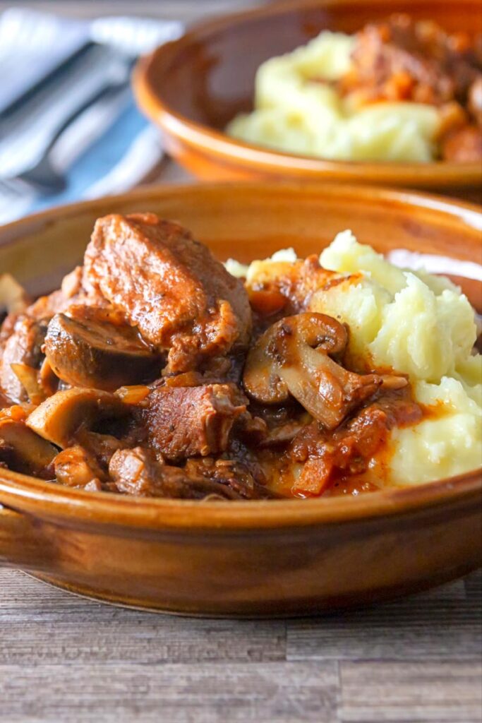 Portrait image of a tomato based wild boar stew with mushrooms served in a an earthenware bowl with mashed potatoes