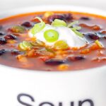 Portrait close up image of a vegetarian black bean soup served in a white bowl with sour cream and green onions with text overlay