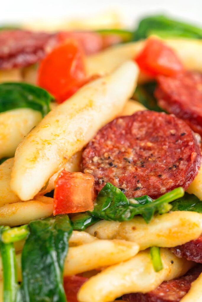Portrait close up image of cured sausage pasta served with wilted spinach and fresh tomato cubes in a white bowl