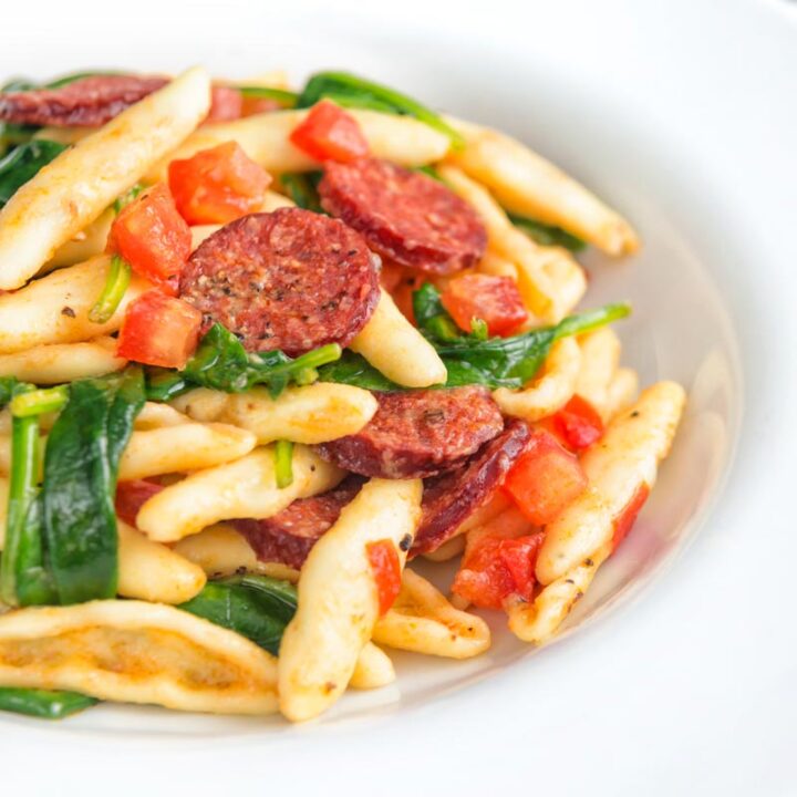 Square image of chorizo pasta served with wilted spinach and fresh tomato cubes
