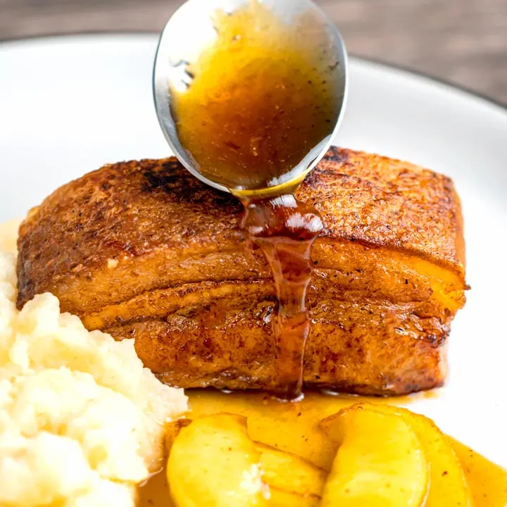 Square image of a sauce being poured from a spoon over Instant Pot Pork Belly served with apples and celeriac mash