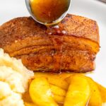Portrait image of a sauce being poured from a spoon over Instant Pot Pork Belly served with apples and celeriac mash