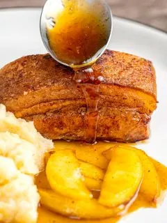 Portrait image of a sauce being poured from a spoon over Instant Pot Pork Belly served with apples and celeriac mash