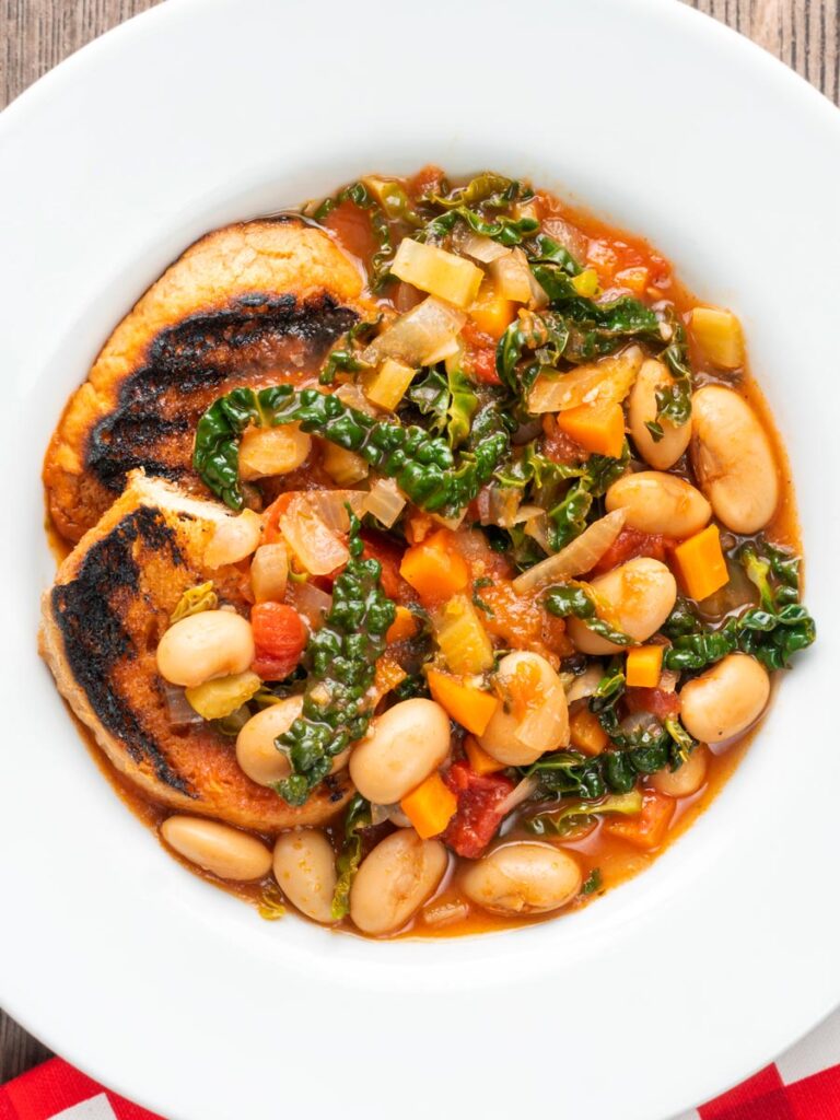 Portrait Overhead image of an Italian Ribollita stew featuring beans, cabbage, carrots, tomatoes and grilled bread