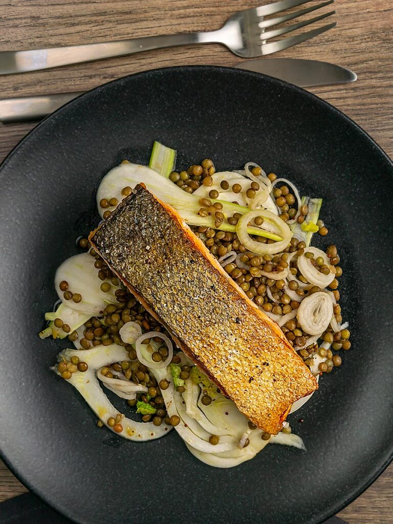 Portrait overhead image of a crispy skinned pan seared salmon fillet served on a black plate with lentils and fennel