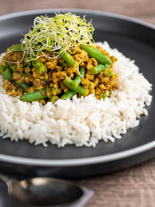 Portrait image of a pork mince curry with green beans served with boiled rice and sprouted onion seeds on a black plate